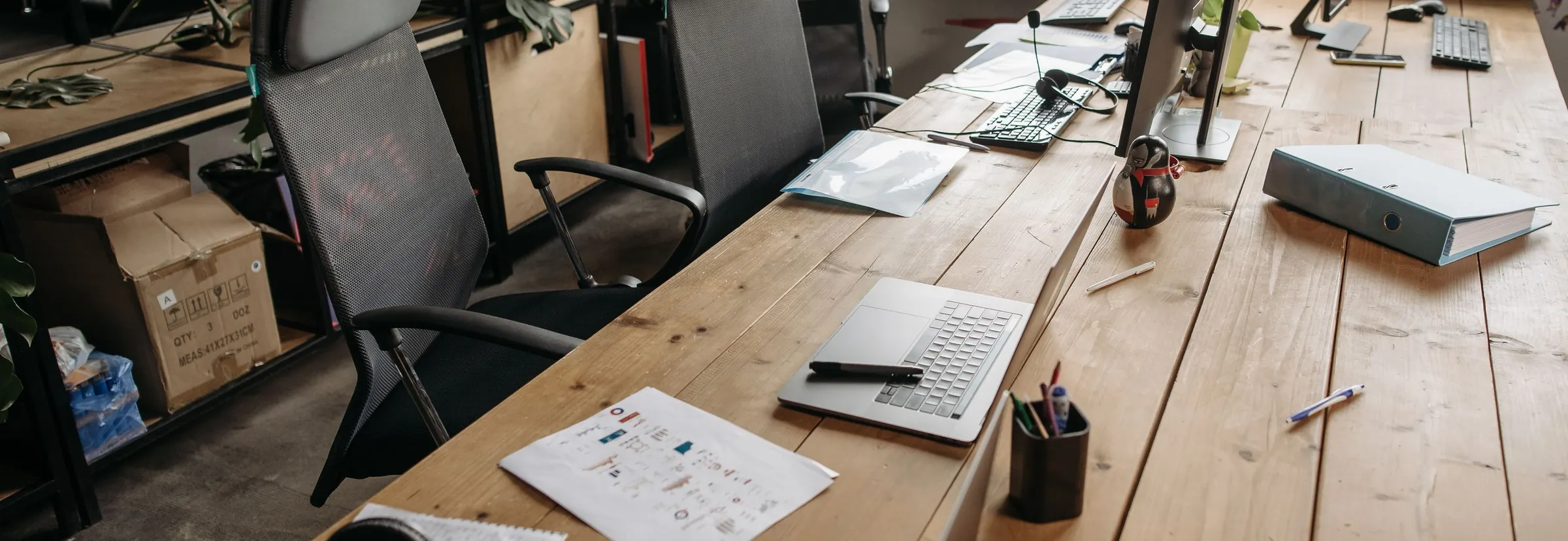 An empty office desk of light wood with a laptop, a printed chart and a binder beside black mesh chairs