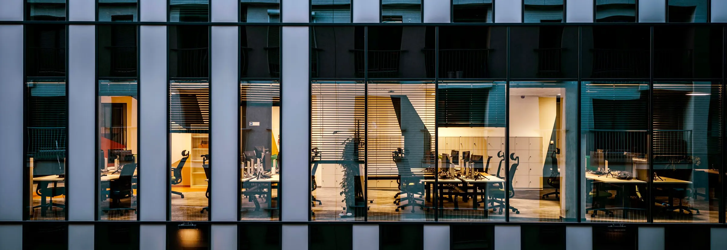 An office building at night, seen through its windows: rows of empty desks and chairs under the lights