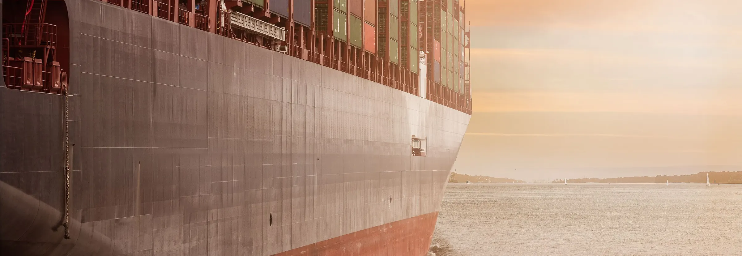 The hull of a loaded container ship at sea, with a coastline on the horizon at sunset
