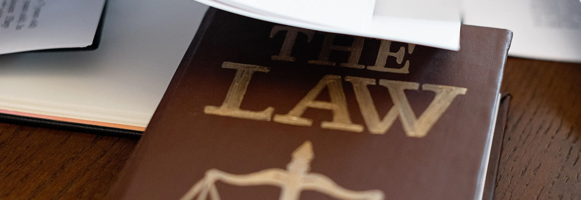 A brown book titled The Law, with scales of justice on the cover, under loose papers on a wooden desk
