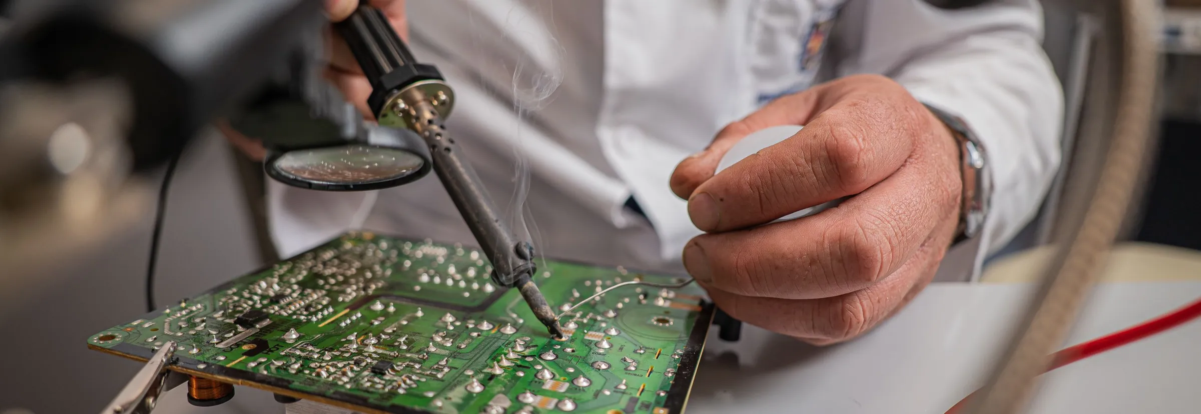 A technician in a white lab coat solders a green circuit board held in a clamp under a magnifier lamp