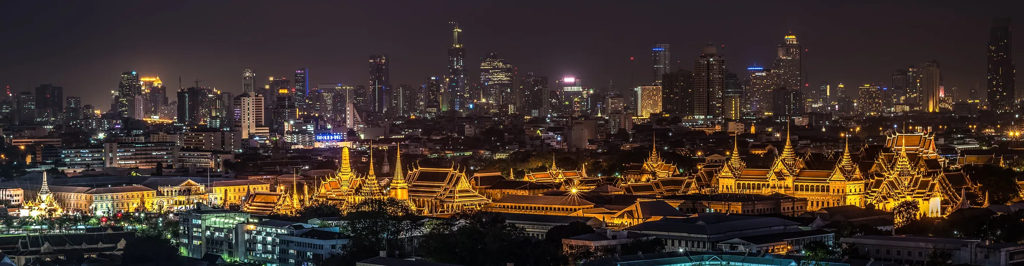 The Grand Palace lit up at night, with the Bangkok skyline behind it