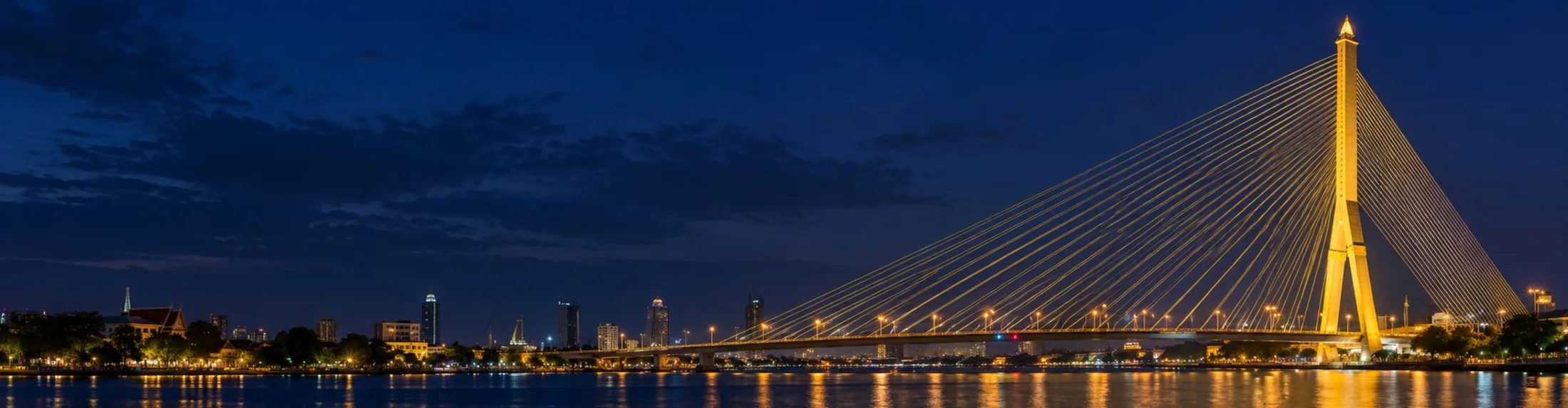 The Rama VIII Bridge lit up at night over the Chao Phraya River, with the Bangkok skyline along the bank